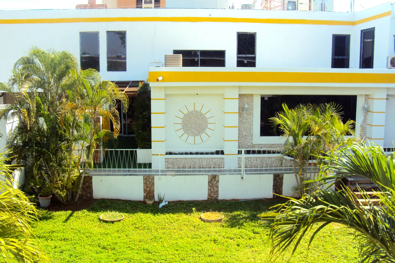 Fachada blanca del hotel con su franja amarilla y el jardín